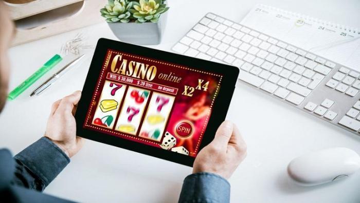 Händer som håller surfplatta med casinospel mot en bakgrund av ett tangentbord, en datormus, en växt, pennor med mera